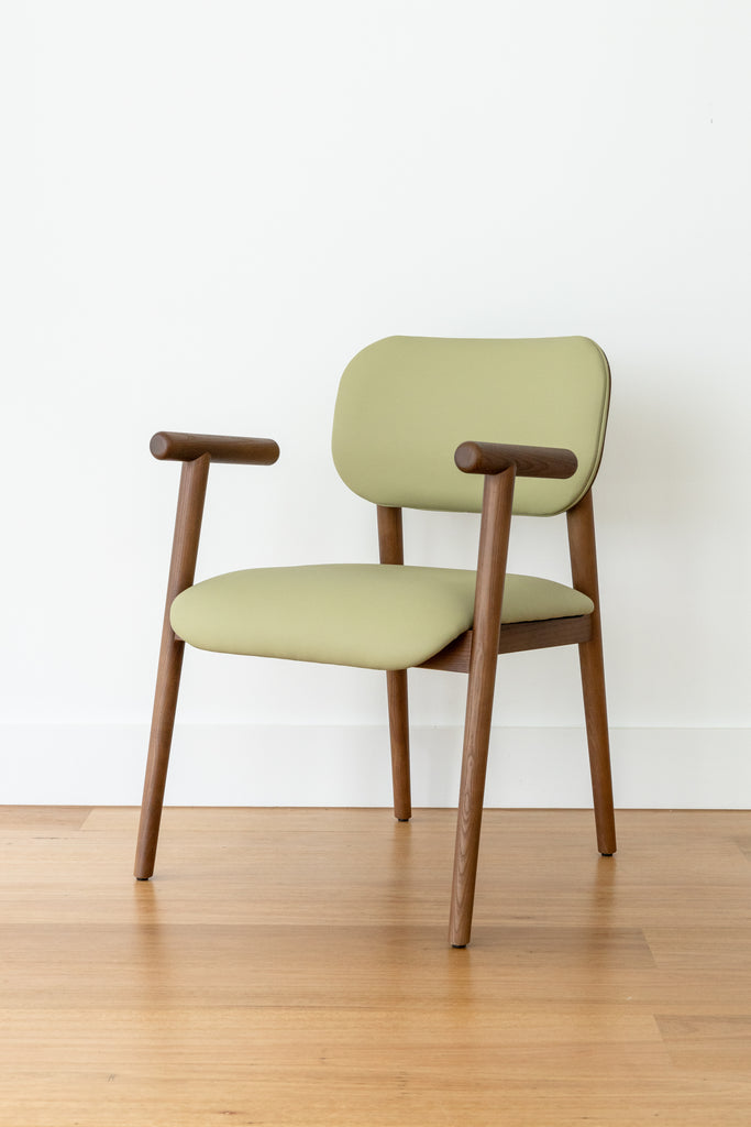 A Kyoto Dining Chair with a green cushioned seat and backrest, solid ash timber legs, positioned on a wooden floor against a white wall.