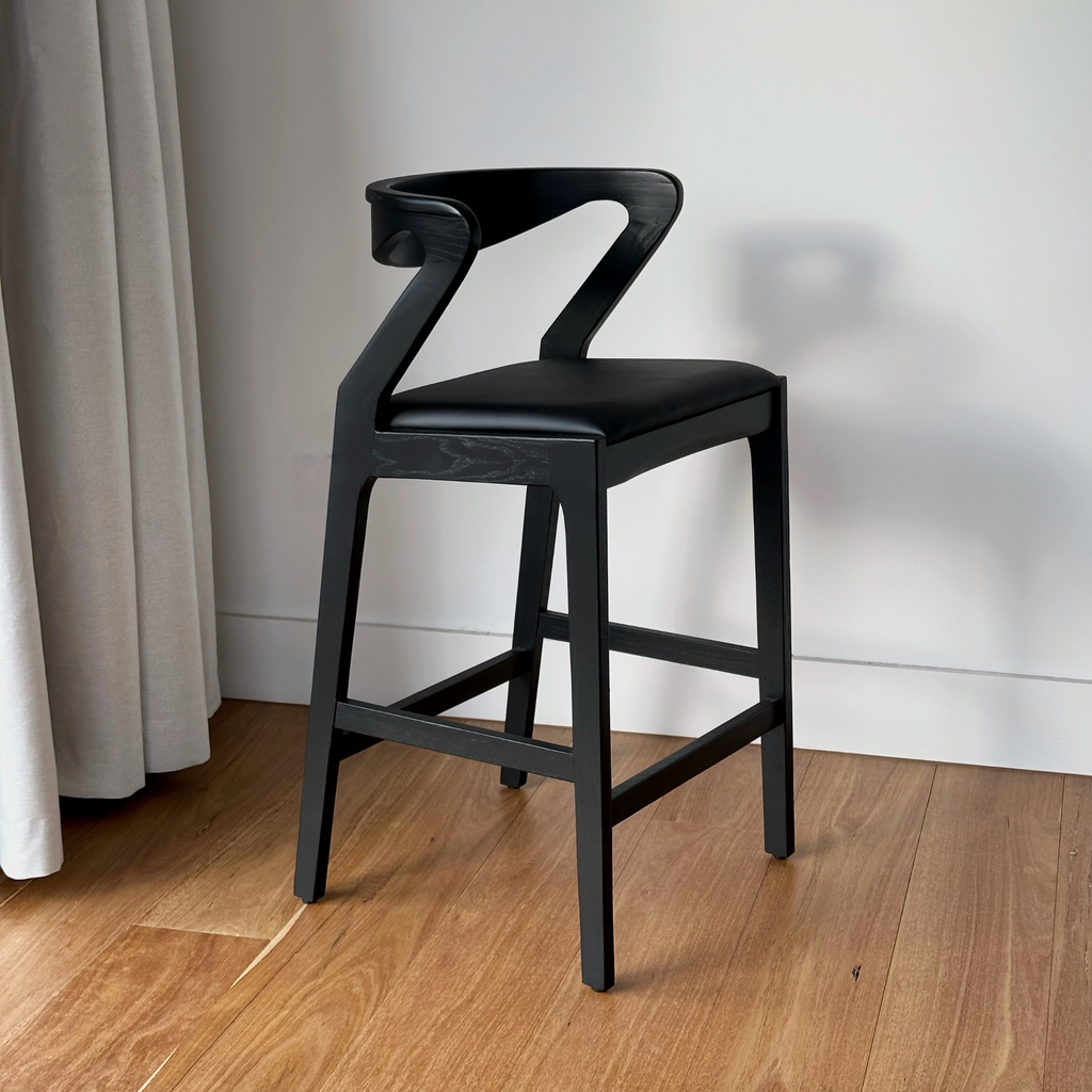 A charcoal timber bar stool with a black cushioned seat, positioned on a hardwood floor against a white wall.
