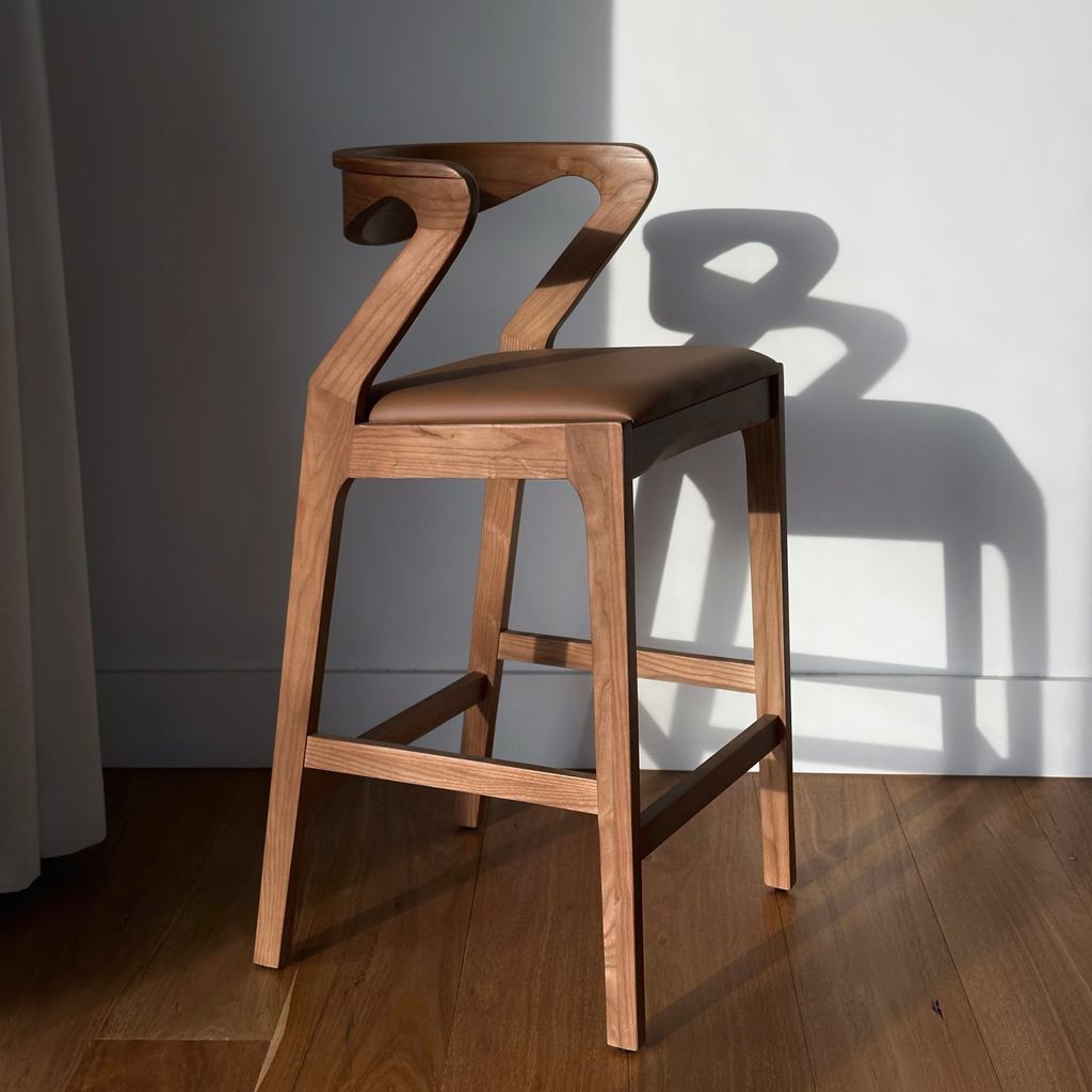 A walnut timber bar stool with a mocha coloured seat cushion, positioned on a hardwood floor with a shadow cast on the wall behind it.